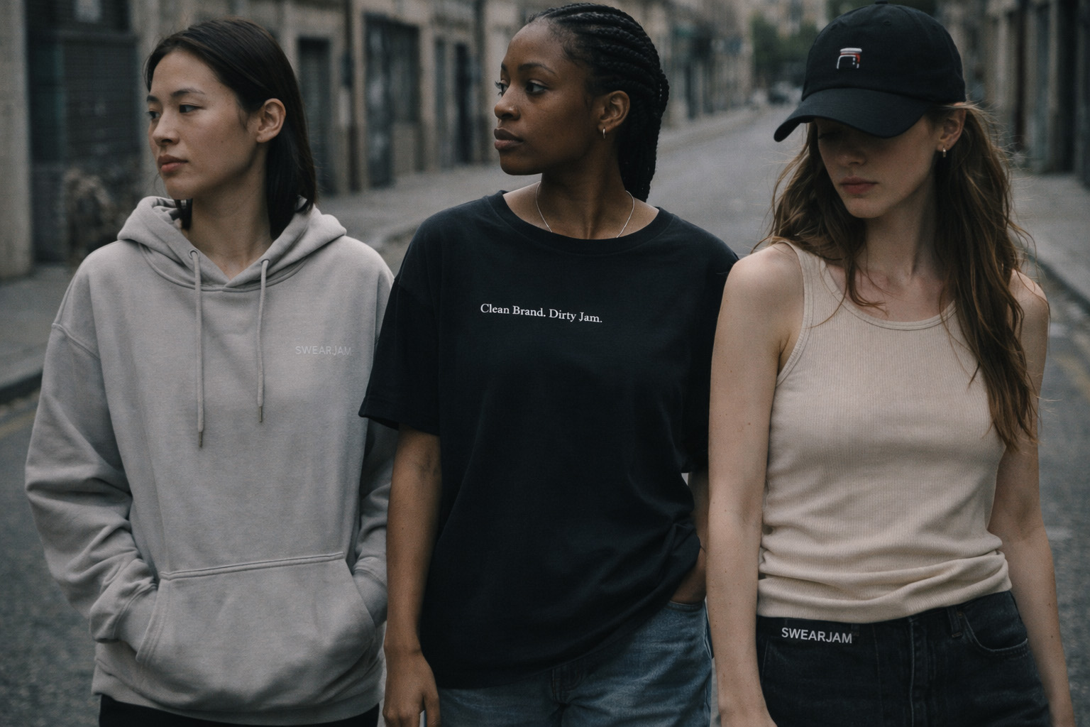 Swearjam Series 001 — three wearers in hoodie, tee, and cap, photographed in low evening light on a cobbled street